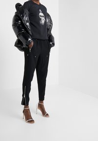 Chaqueta de plumas negra con acabado brillante, sudadera gráfica y pantalones jogger negros ajustados con detalles con cremallera, combinados con sandalias de tacón transparente.