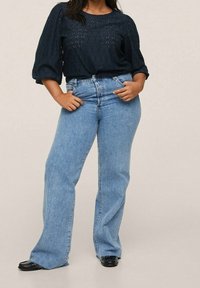 Une femme porte un chemisier noir texturé à manches bouffantes trois-quarts, un jean bleu clair taille haute à jambes larges, et des chaussures noires à bouts carrés.