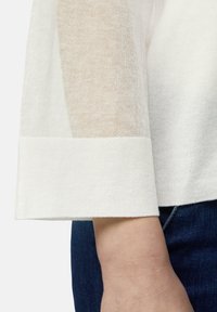 Lekki sweter z kremowej wełny z przezroczystymi beżowymi wstawkami na rękawach, z szerokim mankietem i miękką fakturą. Nosi się go na dżinsy.