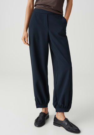Vrouw draagt donkerblauwe taps toelopende broek met elastische manchetten en zwarte loafers, staand met één hand in de zak tegen een effen achtergrond.