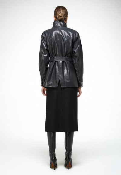 Vrouw met een zwarte leren jas met ceintuur, een zwarte midi-rok en zwarte, kniehoge laarzen met hak, staand tegen een witte achtergrond.