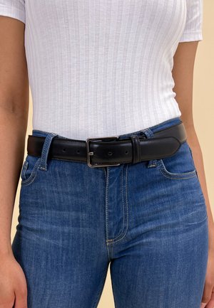 Black leather belt with a rectangular silver buckle, worn over a white ribbed top and blue jeans. Smooth texture and adjustable fit.