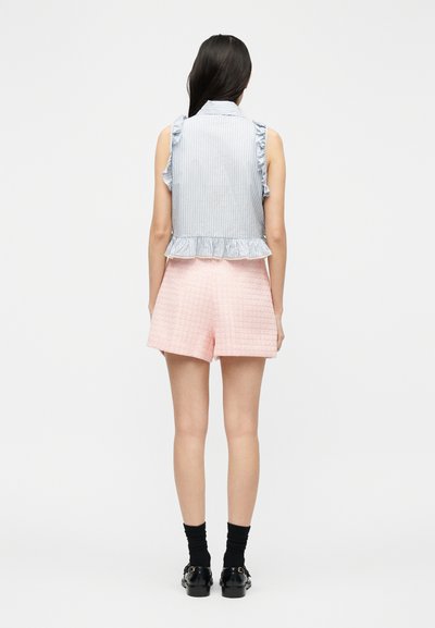 Γυναίκα με μακριά καστανά μαλλιά που φορά μπλε ριγωτή μπλούζα χωρίς μανίκια με βολάν, ροζ κοντά παντελόνια με υφή, μαύρες κάλτσες και μαύρα παπούτσια, με την πλάτη προς το μέρος μας.