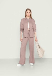 Conjunto de roupa de lazer castanho claro, composto por uma camisa de botão e calças com cordão, combinado com ténis brancos e uma mala de lona bege. Textura suave.