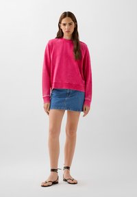 Felpa fucsia con collo a giro e polsini a coste, abbinata a una mini gonna di jeans e sandali neri con cinturini, con suola piatta.