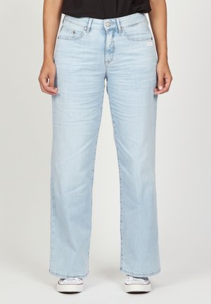 Personne portant un jean droit bleu clair et des baskets blanches, debout devant un fond clair uni.