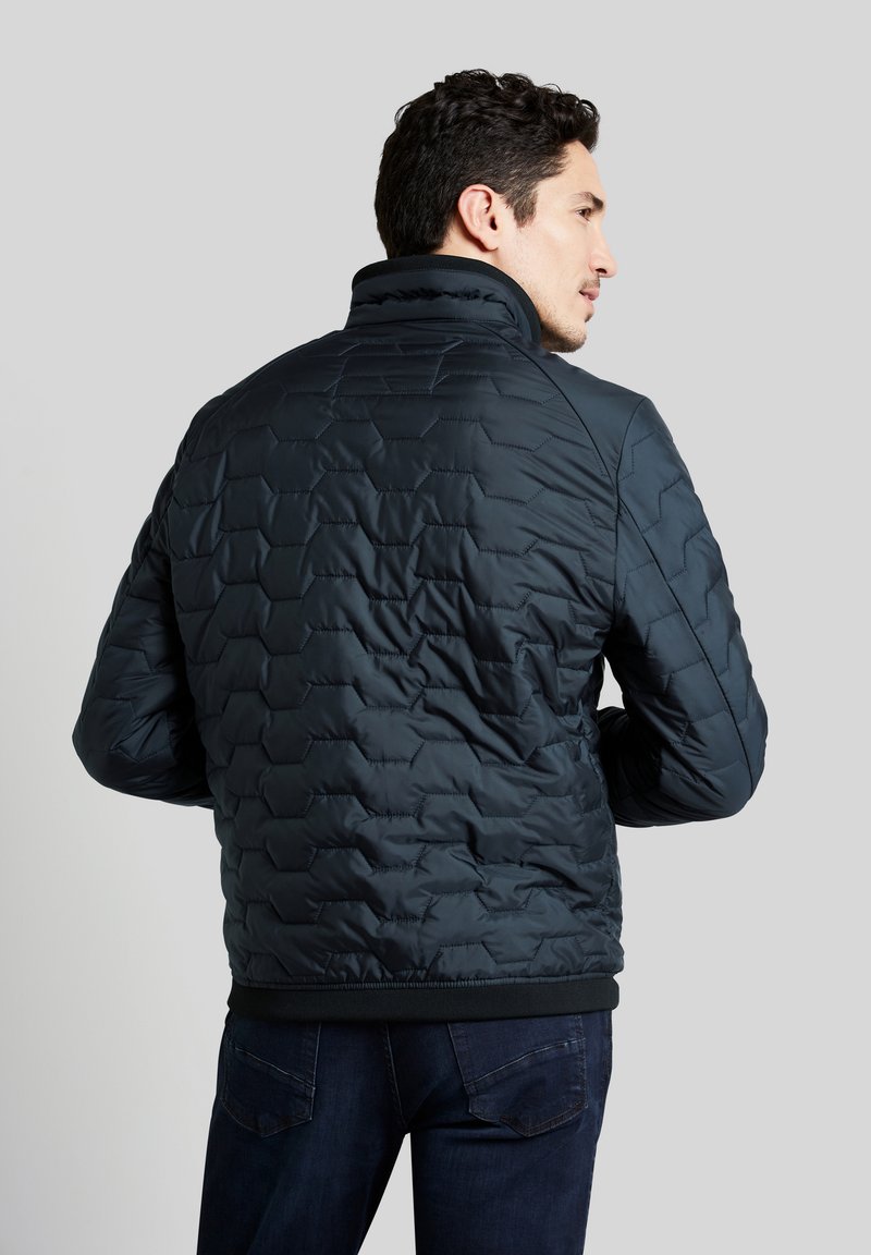 Veste matelassée bleu foncé avec un col haut et un ourlet côtelé. Présente un motif hexagonal et une texture lisse. Portée avec un jean foncé.