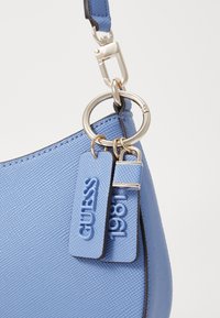 Coin de sac à main texturé bleu clair avec anneau en argent et deux étiquettes indiquant « GUESS » et « 1981 » attachées à une sangle bleue.