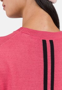 Różowy sweter z dzianiny z prążkowanym kołnierzem, z dwiema pionowymi czarnymi paskami wzdłuż pleców. Gładka faktura i dopasowany krój.