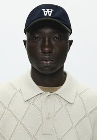 Sötétkék sapka fehér "W" logóval, krém színű, texturált, gyémántmintás póló, bézs aláöltözet látható a gallérnál. Tiszta, minimalista háttér.