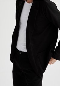 Svart herringbone-blazer med en enkel knappstängning, bärs över en vit t-shirt. Svarta byxor synliga, med en framficka.