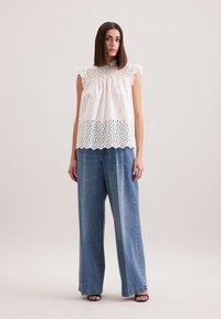 Top bianco in cotone con maniche a volant, dettagli tagliati al laser e orlo ondulato, abbinato a jeans in denim blu a gamba larga. Sandali neri semplici.
