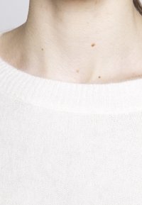 Lekki, kremowy sweter z prążkowanym dekoltem i miękką fakturą, z widocznymi detalami przeszyć i luźnym krojem.