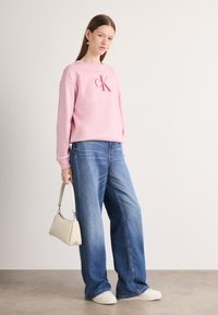 Felpa rosa chiaro con logo "CK", abbinata a jeans in denim blu a gamba larga. Accessoriata con una borsa beige e sneakers bianche.