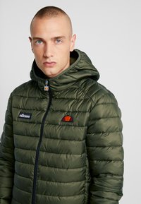 Jachetă puffer verde cu închidere prin fermoar, glugă și design matlasat orizontal. Prezintă accente cu logo brodat pe piept.