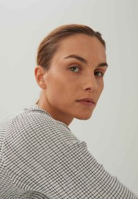 Nahaufnahme einer Frau mit heller Haut, die einen strukturierten, schwarz-weißen Pullover mit Karomuster trägt. Ihr Haar ist zurückgebunden, was einen glatten Teint zur Schau stellt.