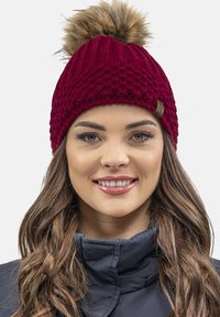 Burgundy knitted beanie with a textured pattern and a pom-pom on top. Features a small leather tag accent.