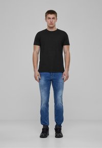 Camiseta de algodón negra, diseño ajustado, combinada con jeans azul desgastados que presentan detalles desgatados y zapatillas negras sobre un fondo liso.