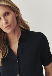 Camisa de manga corta azul marino con diseño de botones, que presenta un cuello con solapas y puños enrollados, fabricada con una tela ligera y texturizada.