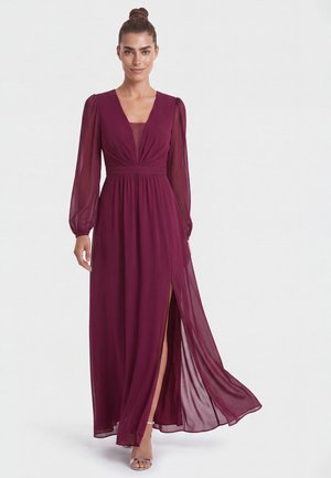 Frau mit Hochsteckfrisur, die ein langes bordeauxrotes Kleid mit transparenten langen Ärmeln und Seitenschlitz trägt, steht lächelnd vor einem einfarbigen Hintergrund.