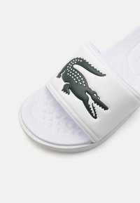 Chausson de bain blanc avec une semelle texturée et une large bande arborant un logo crocodile vert. Matériau synthétique lisse.