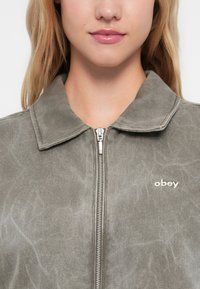 Siva jakna na zadrgo s uskim ovratnikom, ki ima subtilen teksturiran vzorec in bel logo "obey" na prsih.