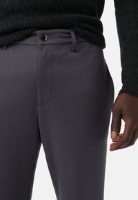 Grijze, gestructureerde broek met een gladde afwerking, voorzien van een sluiting met een knoop aan de voorkant en zijzakken, deels bedekt door een donkere trui.