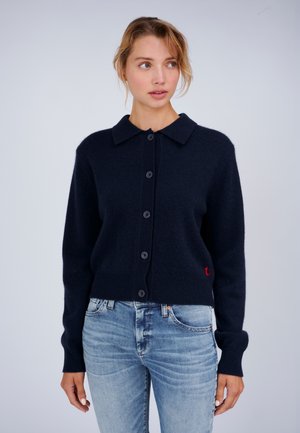Frau trägt einen schwarzen, geknöpften Cardigan mit Kragen und hellblaue Jeans, steht vor einem einfarbigen hellgrauen Hintergrund.