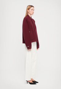 Bordeaux oversized jas met hoge kraag, gestoffeerd materiaal, met ritszakken, gecombineerd met witte wijde broek en zwarte hakken.