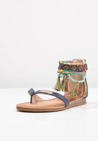 Ploché sandály s kovovou námořnickou modrou sponou, vysoké po kotník s barevnými tkanými detaily a třásněmi, opatřené polstrovanou stélkou a gumovou podrážkou.