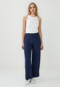 Marineblauwe wijde broek met hoge taille, gecombineerd met een witte mouwloze top. Het model draagt witte sneakers en staat tegen een effen achtergrond.