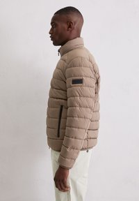 Beige gewatteerd jack met doorgestikt patroon, hoge kraag en met rits afsluitbare voorzakken. Heeft een zwart logoplaatje op de linkermouw.