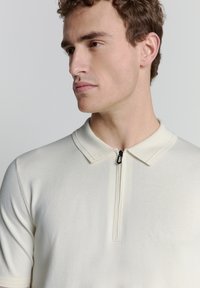 Gebreide poloshirt in off-white met korte mouwen, een ritskraag en een gestructureerde stof. Kenmerkt zich door een subtiele glans en strakke lijnen.