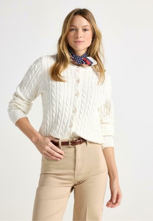 Kvinde iført en hvid kabelstrikket cardigan med knapper, beige bukser med brunt bælte og et marineblåt halstørklæde med prikker og striber.