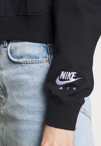 Svart sweatshirt med ribbade muddväskor, med en vit och lila broderad Nike-logotyp. Bärs med ljusblå denimjeans.