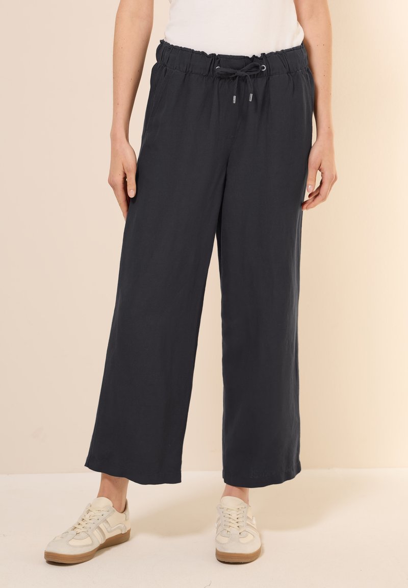 Donkergrijze, lichte, wijde broek met een elastische tailleband en trekkoord. Gladde textuur met een ontspannen pasvorm, gecombineerd met casual sneakers.