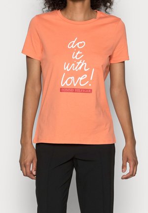 Vrouw draagt een perzikkleurig T-shirt met de tekst "do it with love!" en zwarte broek, beeld bijgesneden van net onder de schouders tot boven de dijen.