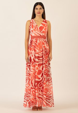 Frau trägt ein ärmelloses, bodenlanges Kleid mit rotem und weißem abstraktem Tiermuster, steht aufrecht mit entspannten Armen an den Seiten.