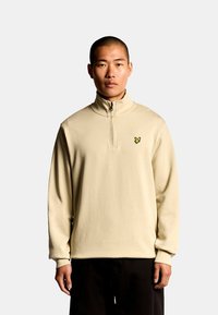 Beige Sweatshirt mit Reißverschluss, hohem Kragen und langen Ärmeln, mit einem kleinen gelben, gestickten Logo auf der Brust. Weicher, strukturierter Stoff.