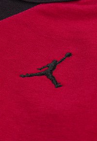 Un tissu rouge avec une surface texturée présente un logo Jumpman noir brodé. Le design comprend un col noir contrastant.