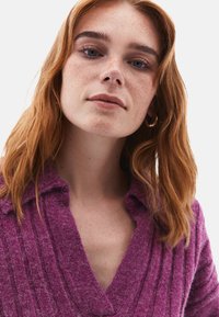 Paarse gebreide top met v-hals, voorzien van verticale ribbels, een gestructureerde stof en een relaxte kraag. Het model heeft roodachtig haar en lichte sproeten.