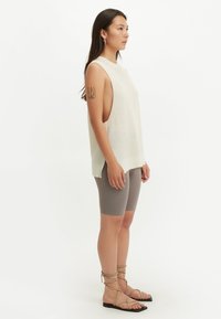 Kvinna klädd i en ärmlös beige nät-topp, taupefärgade cykelshorts och platta sandaler med remmar, stående i profil mot en vit bakgrund.