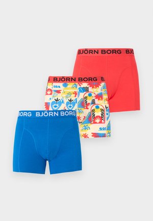 Άνδρας χωρίς πουκάμισο, όρθιος ξυπόλυτος σε φωτεινό χαλί, φοράει μπλε μποξεράκια Björn Borg μπροστά σε απλό φωτεινό τοίχο.