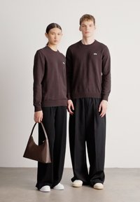 Dois modelos usam sweatshirts castanho-escuros com punhos canelados, combinados com calças largas pretas. A modelo feminina segura uma bolsa castanha. Calçados brancos são usados.