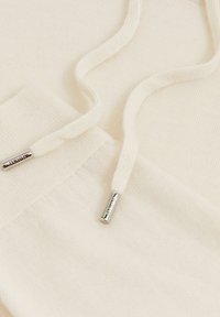 Cremfarbener Hoodie-Stoff mit strukturiertem Finish, ausgestattet mit passenden Kordeln und silbernen Metallenden, die mit "STYLES" geprägt sind.