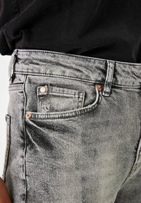 Jean en denim gris avec un lavage décoloré, comportant plusieurs poches, une petite poche avant et des accents en métal cuivré.