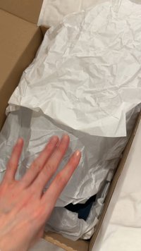 Käsi nostaa rypistynyttä valkoista nenäliinaa ruskean pahvilaatikon sisältä, paljastaen osittain alla olevaa tummaa kangasta.