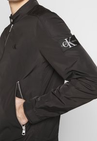 Svart nylon bomberjacka med hög krage, dragkedja fram och sidofickor; har en logopatch på vänster ärm och texturerade detaljer på axlarna.