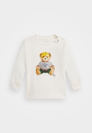 Langarmige crèmekleurige katoenen shirt met een berenafbeelding in een gele pet, grijze sweatshirt en groene shorts; knoopaccenten op de schouder.