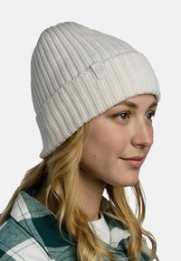 Young woman with long blonde hair wearing a ribbed cream knit beanie and a green and white plaid shirt, facing right with a neutral background.